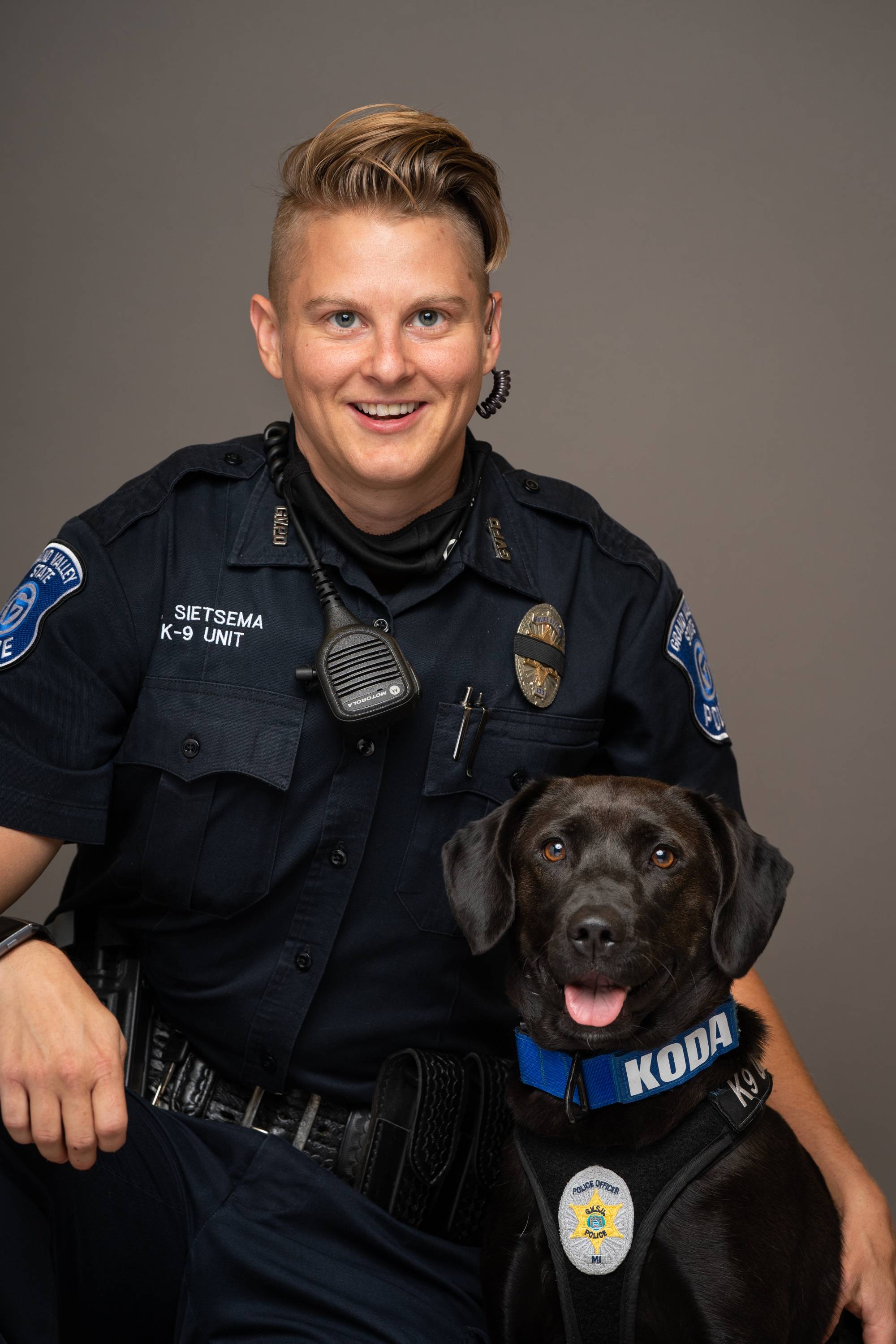 Officer Sietsema portrait photo while wearing police uniform with police dog Koda, who is a black Labrador wearing a harness with a police badge